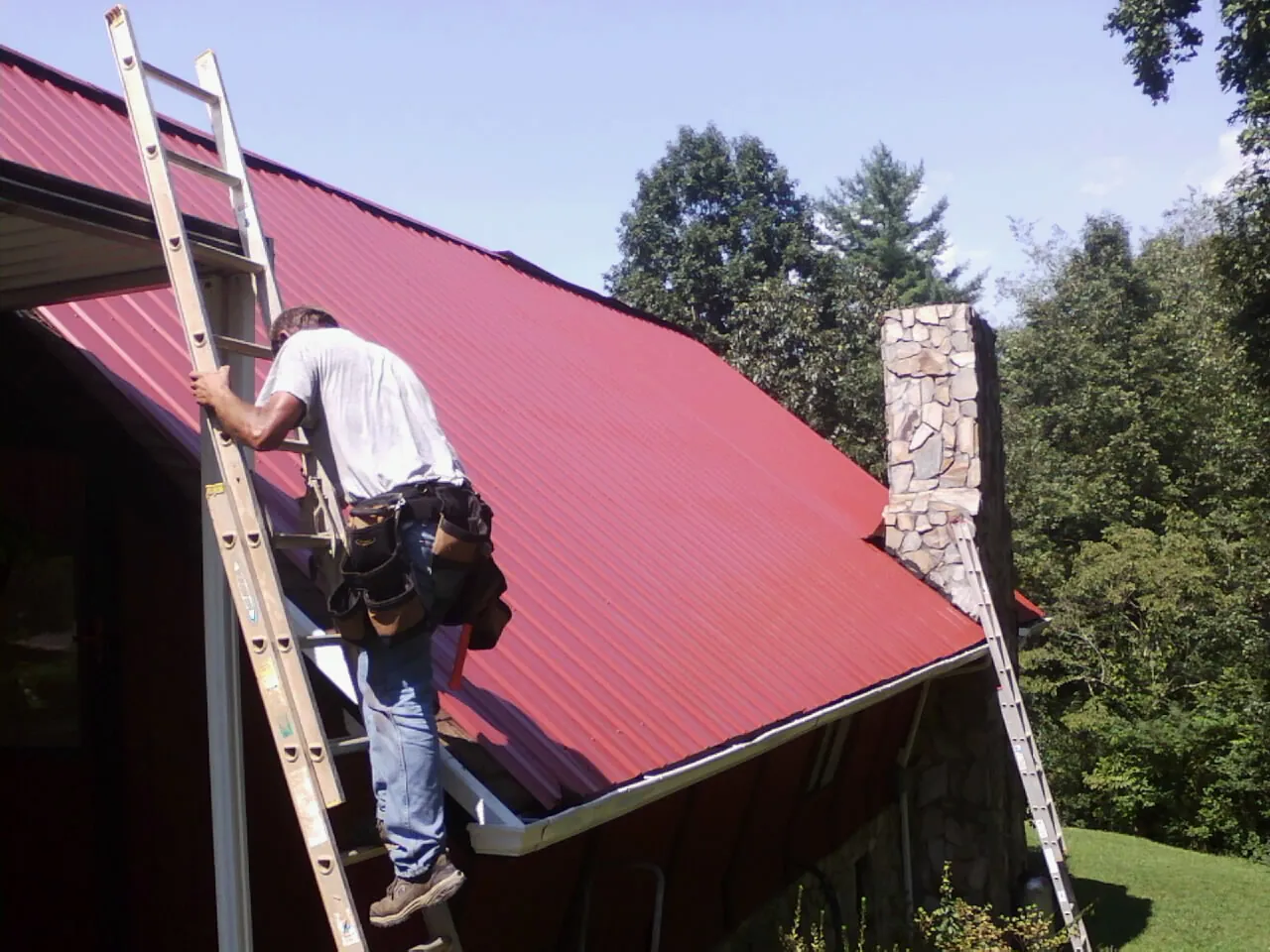 Roofer on a ladder beside a red metal roof and stone chimney — Soffit & Fascia Repair by Roofing Brothers in Burkittsville